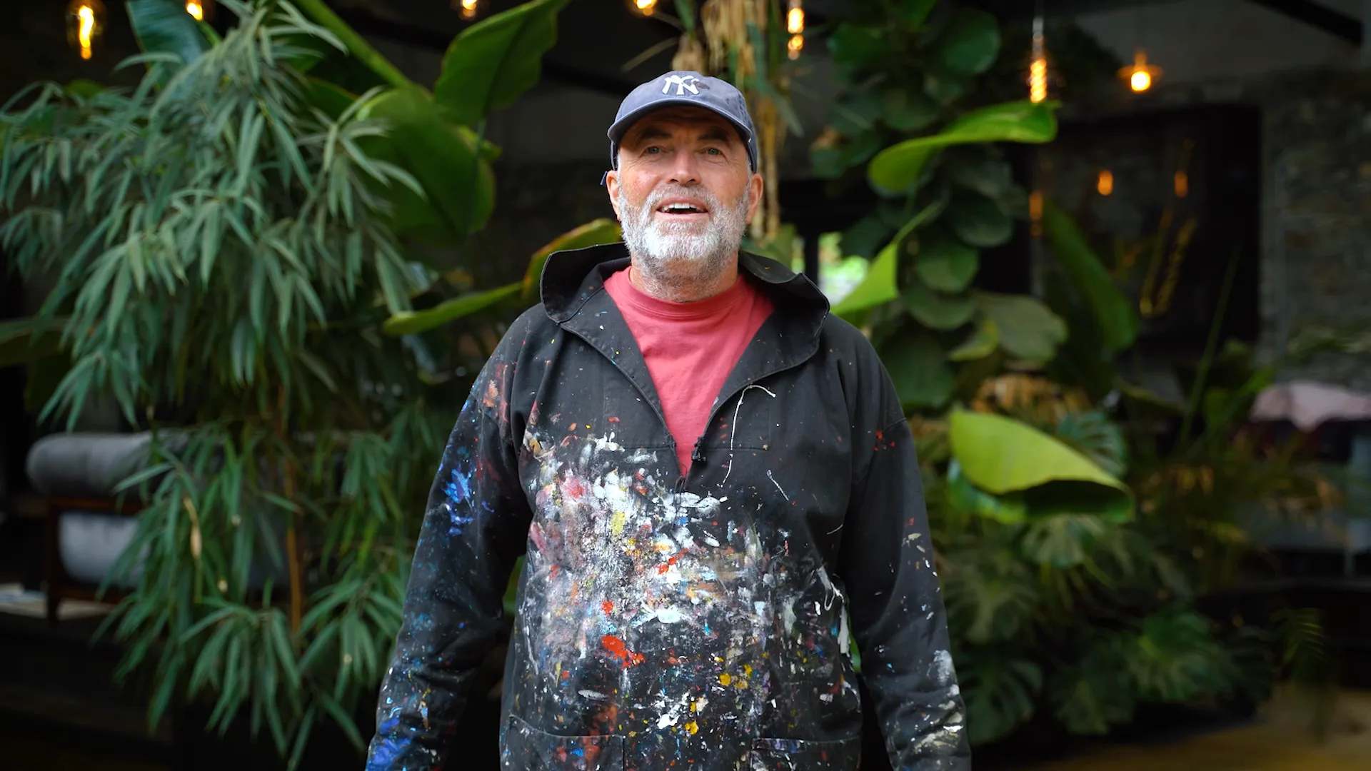 Paul Kenton in a paint-spattered smock and cap, talking among tropical plants