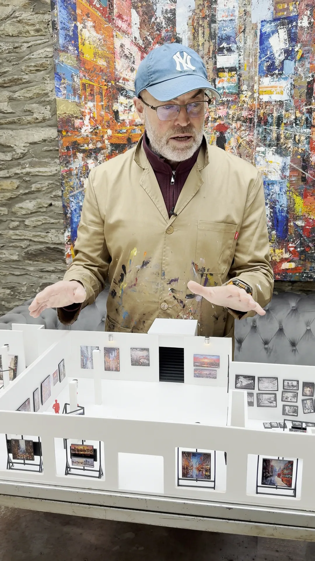 Paul Kenton in a paint-spattered studio coat behind a white scale model of the exhibition, with tiny paintings on its walls